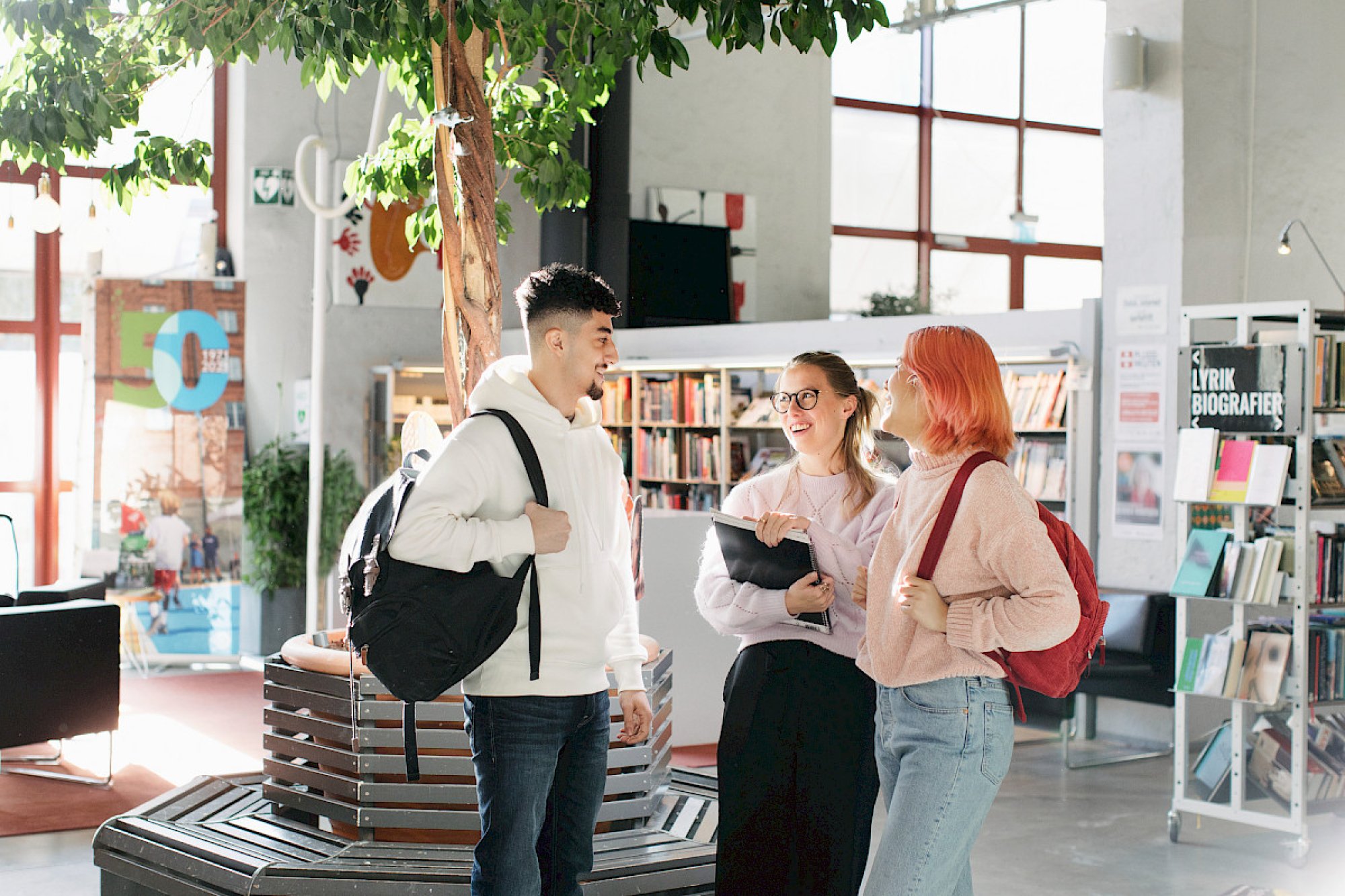 Tre studenter i et bibliotek står i en gruppe og snakker sammen