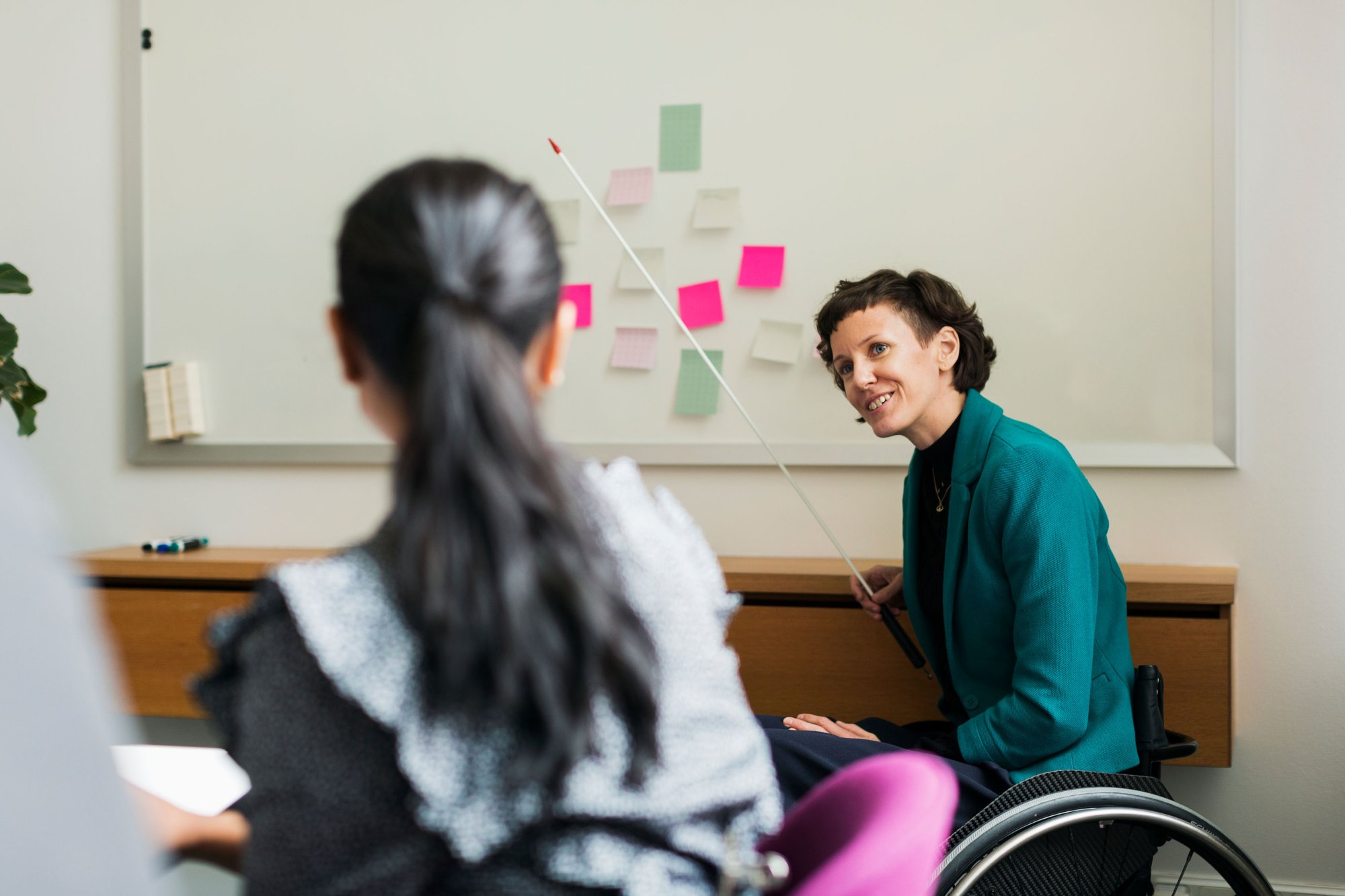 En dame i rullestol sitter foran en tavle med post-it lapper i et klasserom