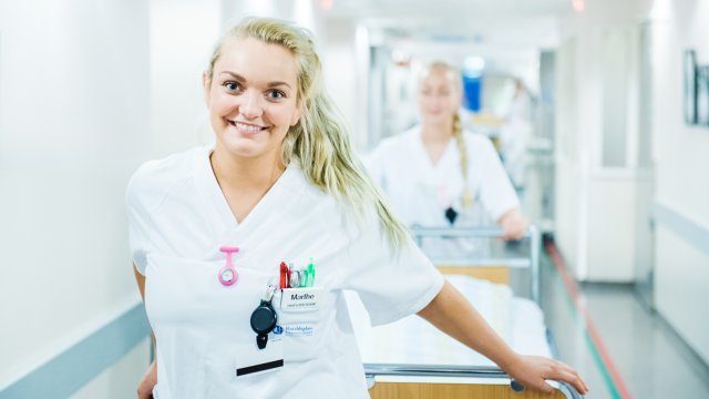 En smilende sykepleier triller seng i sykehuskorridor