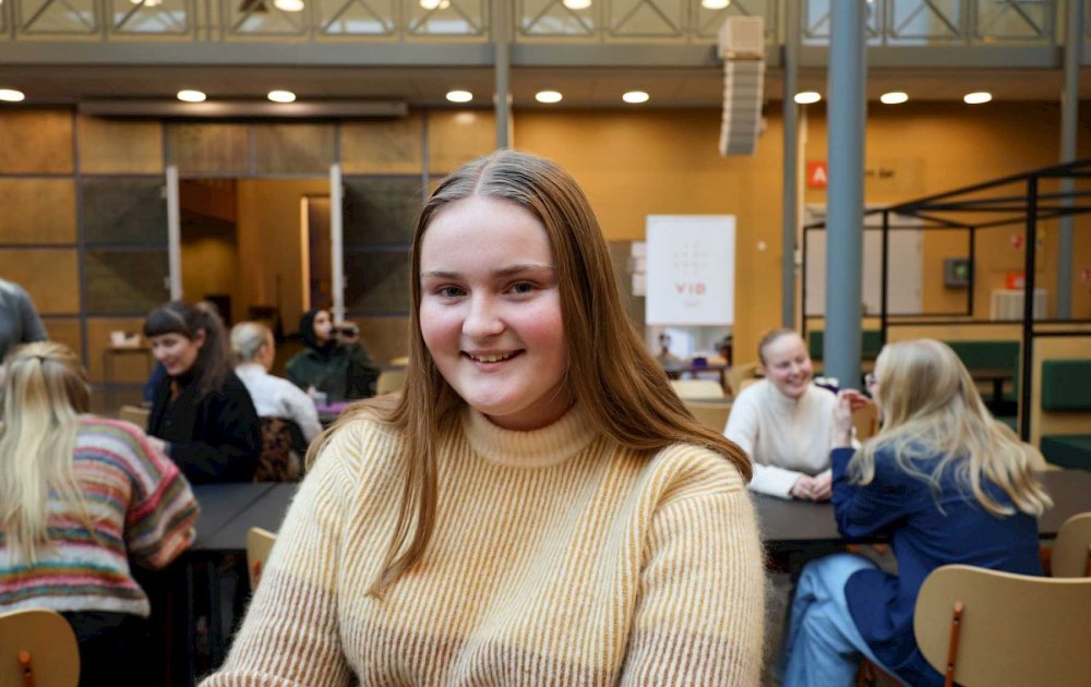 Ung jente ser i kamera og smiler. Bak henne sitter masse studenter ved bord og snakker sammen,