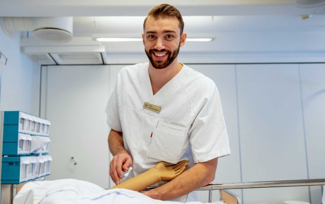 Ung mann med skjegg står foran en sykeseng med en dukke